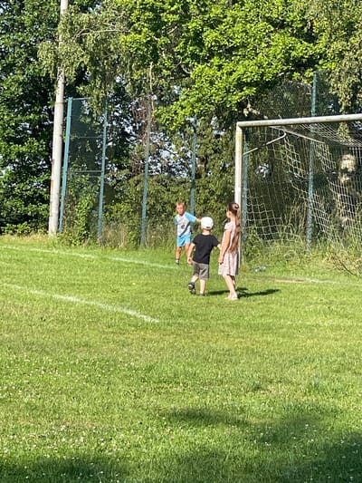 The image depicts a grassy field with a soccer goal in the background. Three children are visible on the field. A boy wearing a light blue shirt with the number "10" and dark shorts stands in the mid-ground. In the foreground, a boy in a black shirt and grey shorts, wearing a white cap, is walking away from the camera. To his right, a girl in a light-colored floral dress with long brown hair is also walking away from the camera. A soccer net is attached to the goalpost. Trees and a fence are visible behind the goal. The scene appears to be outdoors during daylight. The location is Beltershain, Germany.