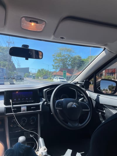 The image shows the interior of a car, likely a Mitsubishi Xpander, as evidenced by the steering wheel emblem. The view is from the passenger seat, looking forward through the windshield. The dashboard is visible, featuring a touchscreen infotainment system displaying the time as 1:16 AM. A USB cable is plugged into the system and dangles down. The steering wheel, gear shifter, and climate control knobs are also in view. Outside the car, a sunny day is depicted with a road, other vehicles, trees, and buildings, suggesting an urban or suburban environment. The location is identified as Ketapang, Indonesia. There are no people clearly visible in the frame. The overall scene is a snapshot of a moment inside a parked or moving vehicle on a bright day in Ketapang, Indonesia.