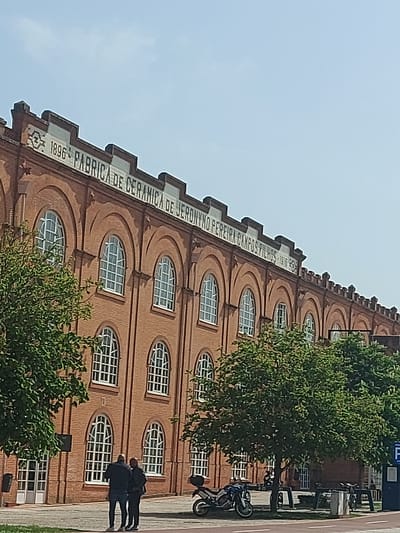 The image shows the Fábrica de Cerâmica Jerónimo Pereira Campos in Aveiro, Portugal. The brick building has rows of arched windows. Text on the building reads "1896 FABRICA DE CERAMICA DE JERONIMO PEREIRA CAMPOS FILHOS 1916-1958." Two people stand in front of the building. A motorcycle is parked nearby. Trees are in the foreground. The sky is visible in the upper left corner.