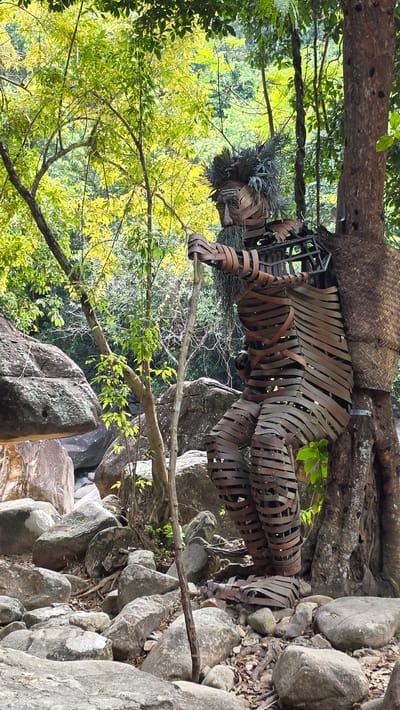 The image shows an outdoor scene featuring a metal sculpture of a human-like figure with a woven basket-like structure above it. The sculpture appears to be integrated into the natural environment, resting on a large tree root and amongst large rocks. The vegetation includes green and yellow-leaved trees. The location is inferred to be in Nam Ninh Hoa, Vietnam, based on the prompt. There are no people visible in the image.