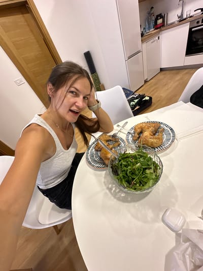 In this image, a young woman is taking a selfie at a round table in what appears to be a modern kitchen or dining area. She is wearing a light-colored tank top and black jeans. Her hair is brown and pulled back. She is smiling and has her hand on her head.

On the table, there are two plates of roasted chicken, a bowl of salad, and a white object that resembles an AirPods case. There are also some crumpled white paper, possibly from takeout packaging. The table is white and appears to be a smooth, reflective surface. The background shows white kitchen cabinets, a wooden floor, and a wooden door. A bottle of water is visible on a countertop in the background.

The lighting suggests it might be daytime. The woman's expression is cheerful and energetic. The setting appears to be a domestic interior, likely a home in Lapčići, Montenegro, given the provided context. There is no visible text in the image.