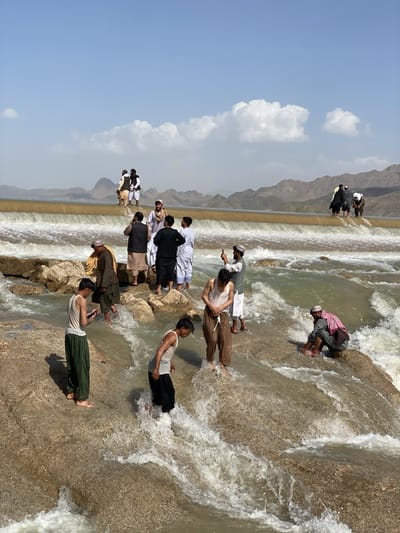 The image shows a group of people at a waterfall in what appears to be a rocky, arid environment. Some people are standing on the edge of the waterfall, while others are in the water. Most are wearing traditional Afghan clothing. A few are using phones to take pictures. The waterfall is flowing over a man-made structure, likely a dam or weir. In the background, there are mountains. There is no indication the location is in the city of Kim, Afghanistan.
