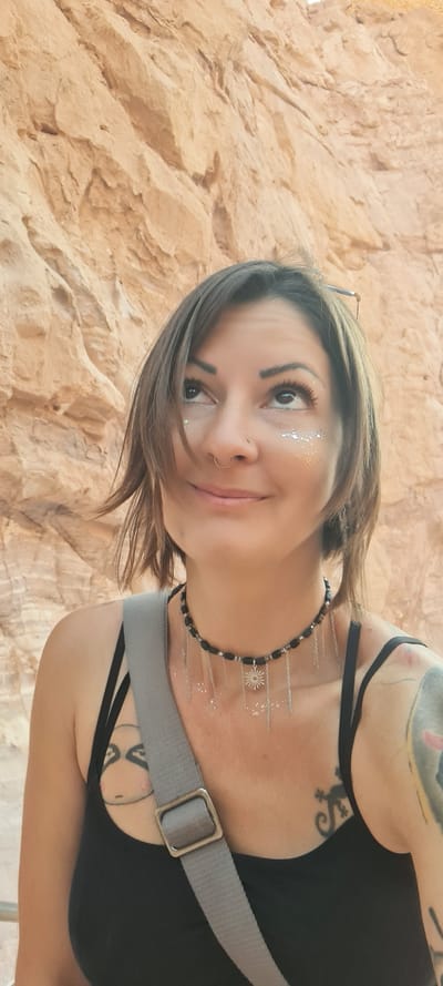 A woman with short brown hair is smiling and looking at the camera. She has glitter under her eyes and a nose ring. She is wearing a black tank top and a black choker necklace. A gray strap is slung across her body. She has tattoos on her arms and chest. The background appears to be a textured rock wall.
The location cannot be pinpointed more specifically within Hevel Eilot Regional Council, Israel, based on the image alone. Further context is needed.