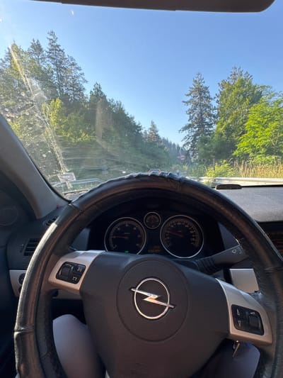 The image is a first-person perspective from inside a car, looking forward. The steering wheel, a black leather-wrapped wheel with silver accents and the Opel logo, is prominently featured in the foreground. The dashboard displays a speedometer and tachometer, with the speedometer needle indicating a speed of approximately 40 km/h. The windshield is visible, showing a clear blue sky and a road winding through a lush, green forest. Trees line both sides of the road, with sunlight filtering through the leaves. A road sign is visible in the distance, suggesting a rural or mountainous area. The overall impression is that of a scenic drive through a wooded landscape. The location is identified as the city of Madan, Bulgaria. There are no people clearly visible in the image.