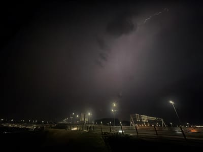 A nighttime photograph captures a dramatic lightning strike illuminating the sky over a coastal area in Badhoevedorp, Netherlands. The image features a dark, overcast sky with a prominent lightning bolt arcing across the upper right quadrant. Below, streetlights line a road or pathway, casting a yellowish glow on the dark, wet ground and adjacent vegetation. In the background, a large, modern structure, possibly a bridge or overpass, is visible, its illuminated panels faintly visible through the haze. The overall scene conveys a sense of a stormy night in an urban or semi-urban environment. There are no people or discernible animals present in the image.