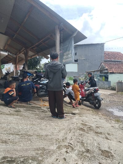 The scene takes place in the city of Kutawaringin, Indonesia, at latitude -6.974 and longitude 107.519. A group of five men are gathered under a corrugated metal roof overhang in front of a building with a faded sign. Several motorcycles are parked in the area. One man is crouched down, seemingly fixing one of the motorcycles, while another stands by the side of the bike. There is a yellow air hose running from the ground towards the bike, and a small yellow air compressor nearby. Two men are standing in the center of the scene, facing the camera, looking at the men working on the motorbike. A young child in yellow clothing stands in front of the motorbike, looking at the men working on it. The ground is covered in a layer of dirt and dust. The sky is cloudy and gray. The man on the right is wearing a blue shirt and black pants, while the other man is wearing a dark gray hoodie. The man crouching down is wearing a blue t-shirt and black pants, and the two men standing are wearing similar attire. The child is wearing a yellow shirt and blue shorts. There is a yellow air hose connected to the front tire of the motorcycle. The man fixing the motorcycle is bending over, with his back to the camera. The man standing next to the bike is facing the camera, but his face is not visible.