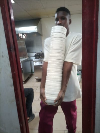 A young Black man is centrally positioned in what appears to be a commercial kitchen in Opete, Nigeria, holding an exceptionally tall and precarious stack of white, round plates. He is wearing a light-colored, short-sleeved shirt and maroon trousers, with his face partially obscured by the impressive column of dishes. His hands are gripping the bottom of the stack, which consists of at least 20 to 25 plates, indicating a task of transporting or organizing dinnerware.
The background features typical kitchen equipment, including stainless steel counters, a commercial cooking range or fryer setup, and a large exhaust hood. The floor is covered with light-colored tiles, and the ceiling shows fluorescent lighting. The scene is framed by dark reddish-brown vertical structures on both sides, possibly door frames, which also cast reflections. On the left side's reflection, the lower leg and foot of another person, wearing dark trousers and a shoe, are faintly visible. The man's expression is focused, highlighting the concentration required to balance the towering stack of plates. There is no visible text in the image.
