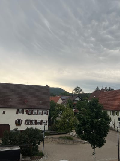 This is an eye-level, medium shot of a European town square in Balingen, Germany. The scene is dominated by a cloudy sky with textured, wispy clouds. In the foreground, a dark rectangular structure is visible on the left. A paved area with hexagonal stones forms the ground, with a lamppost and a street sign indicating a road.

A large, dense green tree stands prominently in the mid-ground, casting a shadow. To the left of the tree, a lighter green tree is visible, and further back, several buildings with red tiled roofs are clustered together. One building on the left is white with brown shutters and multiple windows. Another building to the right has a prominent red tiled roof and white walls with green shutters. In the distance, rolling green hills are visible beneath the cloudy sky, with trees dotting the landscape. The overall impression is of a quiet, overcast day in a European village.