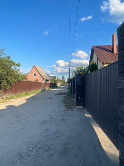 The image depicts a street in the village of Muhawecki, Belarus. The street is unpaved and appears to be a dirt road with scattered gravel. On the left side of the road, there is a brown corrugated metal fence bordering a property with a house behind it. The house has a pinkish-red exterior and a grey roof. Further down the road, more houses and fences are visible. On the right side of the road, there is a tall, dark-colored metal gate and fence, with brick pillars supporting it. An orange safety light is mounted on one of the brick pillars. The sky is blue with scattered white clouds, and the sun is casting shadows on the road. Trees and greenery are present on both sides of the street. The scene appears to be a residential area with a rural or suburban feel.