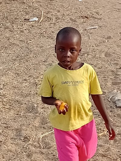 A young child, appearing male, stands in a dry, open area in Nata-Ala, Nigeria. The child has dark skin and short, dark hair, looking directly at the camera with a neutral expression. He wears a light yellow short-sleeved t-shirt with dark lettering, including "DAFFY LUCK" visible on the chest, and bright pink long pants. In his left hand, he holds a small, orange, fruit-like object. The ground surface is composed of dry, light brown soil or sand, sparsely covered with scattered dried plant matter, twigs, and small pieces of white plastic debris. The scene is illuminated by daylight.