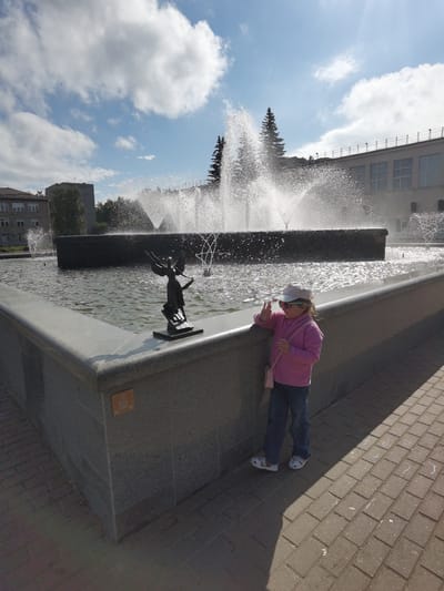 A young girl wearing a pink jacket, blue jeans, a white cap, and sunglasses stands near a large fountain in Chaikovsky, Russia. She is leaning on the stone ledge of the fountain, with her right hand raised in a peace sign and her left hand holding her chin. A small pink purse is slung over her shoulder. The fountain features a dark, rectangular basin with several water jets spraying into the air, creating a shimmering effect. A small, dark bronze statue of an angel stands on the edge of the fountain in the foreground. Behind the fountain, there are buildings and tall evergreen trees. The ground around the fountain is paved with bricks. The sky is blue with scattered white clouds, and the sun is shining, casting shadows.