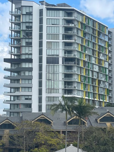 The image shows a modern, multi-story apartment building in Brisbane, Australia. The building features a white facade with balconies and numerous windows. Sections of the building are accented with vertical panels in vibrant colors, including yellow and green. In the foreground, there is a lower building with a dark gray tiled roof and gabled dormer windows, partially obscured by trees. Two palm trees stand in front of the lower building, and other lush greenery is visible. The sky above is partly cloudy with blue sky visible.