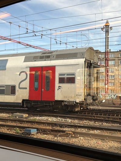 The image presents a detailed view of railway infrastructure and an adjacent urban environment, observed from an interior perspective through a window, located in Saint-Gilles - Sint-Gillis, Belgium.
A light grey and white double-decker railway carriage occupies the midground, positioned on one of several parallel tracks. The carriage displays a large grey numeral "2" on its left side and the identification number "62066" on a white panel below a smaller window to the right of the doors. Two prominent red double-doors with reflective windows are centrally located on the carriage's visible side. The lower section of the carriage, particularly around the doors and wheels, exhibits visible dirt and grime. The right end of the carriage shows coupling mechanisms.
The foreground and midground are dominated by multiple parallel railway tracks, constructed with steel rails resting on a bed of ballast. Various trackside equipment, including a blue rectangular box and a grey square box, are visible between the tracks. Overhead, a dense network of electrical catenary lines and support structures spans the upper portion of the scene, set against a predominantly clear blue sky with faint wisps of clouds.
In the background, a tall, red and white striped utility or signal mast, equipped with lighting elements at its apex, rises above the tracks. Behind this mast and extending across the horizon are several multi-story urban buildings featuring numerous windows. Graffiti is discernible on a lower wall or barrier situated in front of these background structures. No individuals are present in the depicted scene.