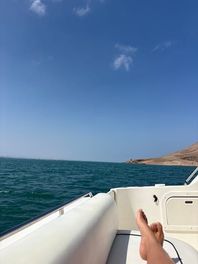 The image depicts a person's bare feet resting on the white cushioned seating of a boat. The boat is on the water, with a clear blue sky above and distant land on the horizon. The land appears to be a hilly, arid coastline. In the far distance, on the left side of the horizon, a city skyline with industrial elements such as cranes and buildings is visible, likely representing a port or urban area. This skyline is consistent with the port facilities of Callao, Peru. The water is a deep teal color, with small waves indicating a gentle movement. The overall scene conveys a sense of relaxation and being at sea.