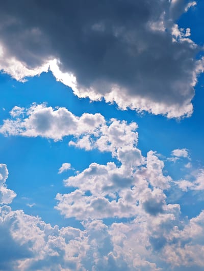 The image depicts a bright blue sky filled with various types of clouds. In the upper portion of the frame, a large, dark gray cloud dominates, with a sliver of bright white light peeking through its lower edge, suggesting the sun is behind it. Below this, numerous white, fluffy cumulus clouds are scattered across the sky. Some of these clouds are large and voluminous, while others are smaller and more wispy. The overall impression is that of a partly cloudy day. The scene is entirely natural, with no man-made structures or living beings visible. The image is a vertical shot looking upwards at the sky.