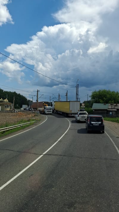 The image depicts a traffic jam at a railway crossing in Сулемка, Russia. The sky is partly cloudy with dramatic white cumulus clouds against a bright blue background. A paved road curves through the scene, marked with white lines. Several vehicles are stopped on the road, including a black minivan in the foreground, a white SUV, a white truck with a yellow trailer, and other trucks further back.
The railway tracks are visible to the left of the road, with a small, green and yellow building adjacent to them. There are utility poles and power lines throughout the scene. On the right side of the road, there are several wooden houses with green roofs and fences, suggesting a rural or semi-rural setting. The general environment appears to be a transitional zone between a populated area and the countryside, with trees lining the background. The overall impression is a snapshot of everyday life interrupted by a common transportation bottleneck at a railway crossing.