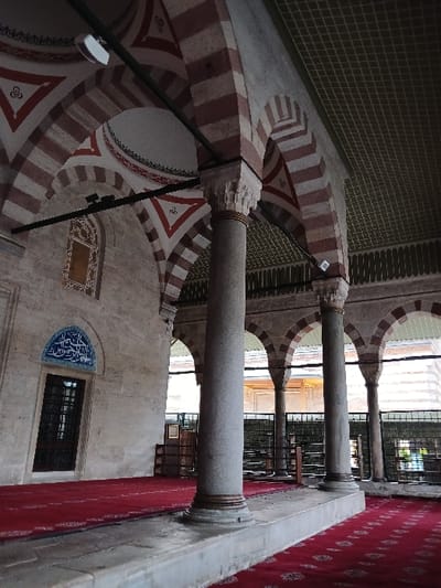 The interior of the Süleymaniye Mosque in İstanbul, Türkiye, located at 41.027 latitude and 28.98 longitude. The image shows a hallway with multiple columns supporting the arched ceiling. The hallway is tiled with a red and white patterned carpet. The walls are made of stone and have a white plaster finish. The ceiling is tiled with green and white squares. The archways are painted with alternating red and white stripes. A window with black bars is visible in the left wall. The mosque is empty of people.