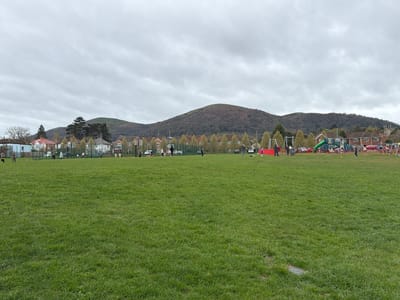 The image depicts a large grassy field in the Malvern Hills, United Kingdom. In the background, a mountain rises behind houses and trees. A playground is visible to the right. Several people are scattered across the field, some playing games and others standing in groups. The sky is overcast.