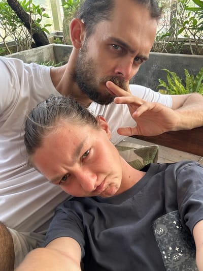 The image is a close-up, angled selfie-style portrait of two individuals, a man and a woman, in what appears to be an outdoor or semi-outdoor setting.
On the left, a man with a dark beard and mustache looks directly into the camera with an intense gaze from his light-colored eyes. He is wearing a plain white t-shirt, and his right hand is raised with fingers near his chin and mouth.
Adjacent to him, on the right, a woman also looks directly into the camera. She has fair skin, rosy cheeks, and light brown eyes, with reddish-blonde hair showing visible grey strands. She is wearing a dark grey t-shirt that features a grey and white digital camouflage patterned pocket on the chest. Her expression is relaxed and engaging.
The background shows lush green tropical foliage and hints of a concrete structure, suggesting a garden, patio, or balcony setting. A wooden surface is visible between the two, and a green and white checkered cushion is partially seen behind the woman. The lighting suggests it is daytime. Both individuals appear to be posing casually for the photograph. There is no visible text in the image.