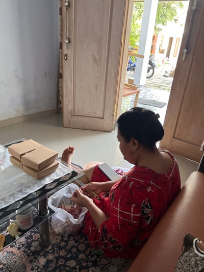 The image depicts an interior scene likely within a residential dwelling in Jambearum, Indonesia. The focus is on a woman, viewed from behind and to her side, wearing a red and white patterned garment. She is seated on the floor, leaning towards a glass-topped coffee table. Her hands are engaged in an activity, appearing to be preparing food, with a plastic bag containing diced meat and a knife visible. The table also holds stacked brown cardboard boxes and a white mug. In the background, a wooden door is ajar, revealing a sunlit outdoor area with a motorcycle and foliage. The floor is tiled, and a patterned rug is visible beneath the table and the woman. The overall impression is one of domestic activity within a home.