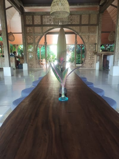 The image depicts the interior of a building in Ubud, Indonesia, characterized by its open-air design and natural elements. A long, dark wood table with a vase containing a flowering plant is the focal point. Cushions line the table's perimeter, indicating seating. The building's architecture incorporates ornate wooden panels and arches, allowing natural light to filter through, highlighting the surrounding lush greenery. In the background, a person is seated at a desk, suggesting an office or workspace. The overall atmosphere is serene and natural.