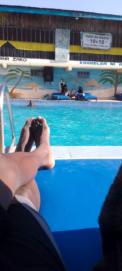 A person relaxes by a swimming pool, feet visible in the foreground. In the background, a mural depicts a beach scene with palm trees, sun umbrellas, and figures sitting on loungers. A building features corrugated metal siding in yellow and black. Painted text in Swahili translates to "Your Choice," "Swimming is a..." and a sign reads, "Waste Awards 10+10 For Fluency and Logic." A "Look Out" sign is also visible.