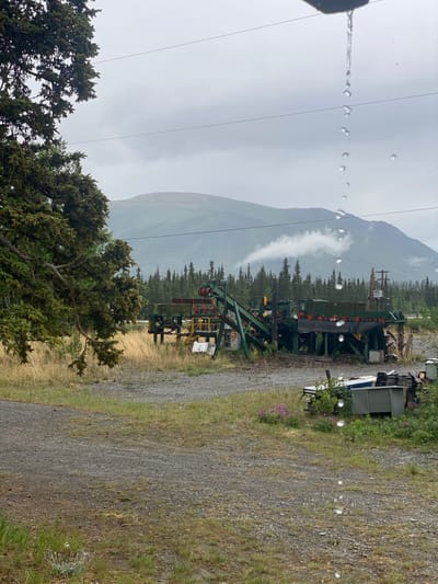 This media file captures a scenic view of a mountainous landscape in Haines Junction, Canada, likely on a cloudy or rainy day, as evidenced by the water droplets visible on the lens in the foreground.
The primary object in the mid-ground is a large, industrial-looking green structure, possibly a conveyor belt system or a section of mining equipment, with a dark belt running along its length and visible mechanical components. It is situated on a gravel path that winds through a grassy and wooded area. In the background, a towering, mist-shrouded mountain dominates the skyline, with a band of clouds or fog clinging to its slopes. Dense evergreen trees form a natural border between the equipment and the mountain.
To the right of the industrial structure, a grey shipping container-like object is visible, suggesting a possible base of operations or storage area. Some scattered purple wildflowers are seen growing in the grass near the gravel path. In the foreground, the tops of dark green coniferous trees are visible, framing the scene and adding to the natural environment. The overall mood is somewhat subdued due to the overcast weather, evoking a sense of rugged natural beauty and industrial presence within a remote setting. No text is visible in the image.