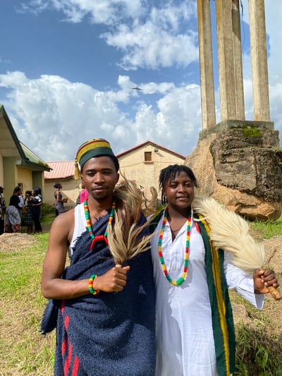 Two individuals, adorned in traditional attire, are captured in a low-angle shot against a backdrop of a cloudy blue sky and lush green grass. The person on the left, wearing a white garment with a green and gold trim, has a white, furry accessory around their neck and is holding a wooden object. Their hair is dark, and they are smiling. The person on the right wears a dark blue garment over a white shirt, with a beaded necklace and bracelet in red, yellow, and green. They are also wearing a colorful cap with a tassel. Both individuals appear to be of African descent.
The setting includes a light-colored building with a red roof and several concrete structures in the background. A bird is visible flying in the sky. The overall atmosphere suggests an outdoor event or gathering, possibly a cultural ceremony or celebration, given the traditional clothing. The lighting indicates it is daytime, with the sun casting shadows, and the presence of clouds suggests variable weather conditions. There is no visible text in the image. The emotions conveyed are positive, with smiles and a relaxed posture. The location context is identified as Wang, Nigeria.