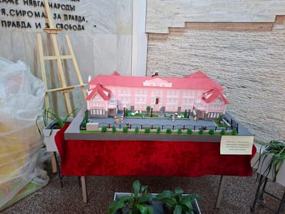 The image shows a detailed architectural model of a pink building with a red roof, adorned with a Bulgarian flag, representing "SU Hristo Botev." This model includes miniature trees, people, and fences, and is displayed on a red-draped table. The model, according to a small sign in Bulgarian, was crafted by students receiving additional support for personal development. The scene is set indoors against a backdrop of stone walls, an easel, and plants in pots. The location is likely in Bulgaria, based on the Cyrillic text and the building's name. "SU Hristo Botev" is the name of a school in the city of Nova Zagora, Bulgaria.