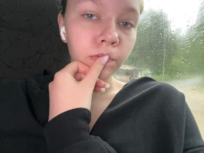 A young woman with light skin and light blue eyes is pictured in a close-up, waist-up shot. She is wearing a black sweatshirt and white earbuds in her left ear. Her right hand is raised, with her index finger touching her lips. Her nails are light pink. In the background, water droplets streak down a window, obscuring a blurry view of trees and a small building, suggesting a rainy day. The scene appears to be indoors, possibly in a vehicle, given the reflection on the window. There is no discernible information in the image to place the location in Krasnokamsk, Russia.