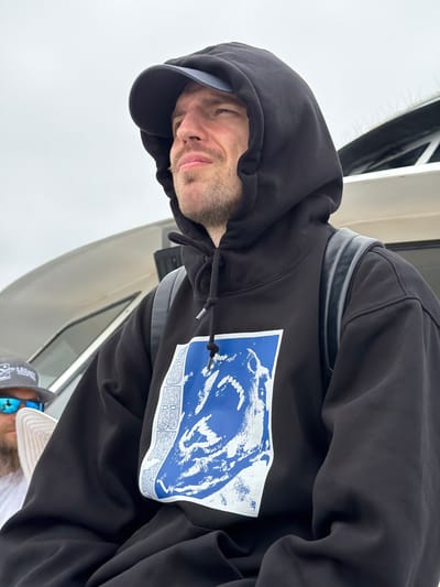 The image, taken from a low angle and originally rotated, presents a close-up portrait of a man. He wears a black hooded sweatshirt with the hood up and a dark baseball cap. His face has short stubble, and he looks towards the viewer with a relaxed, slightly smirking expression.
A prominent graphic adorns the front of his black hoodie. It is a blue rectangular patch featuring a distressed white illustration of a stylized animal head, possibly a wolf or bear, depicted mid-snarl or roar. Below this illustration, faint white text is visible, spelling out "ZOMBOY" and potentially other words like "BASS" or "DUB" with a question mark on the right side. A black drawstring hangs from the hood. A dark strap, possibly from a backpack, is visible over his left shoulder (viewer's right).
Partially visible in the background, positioned below the main subject's arm, is another person wearing a light-colored cap and sunglasses. Text on this cap includes "LEGACY," and a faint "CAT" logo is discernible.
The setting appears to be on a boat or vessel, with white and gray structures visible behind the subjects. The background shows an overcast sky, suggesting a cloudy daytime. The casual attire and marine environment are consistent with a relaxed outdoor or semi-sheltered setting in San Diego, United States.