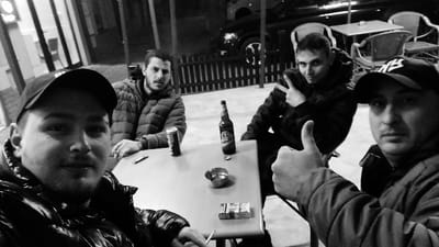 A grayscale photograph depicts four adult males seated around a square table on an outdoor terrace in Senec, Slovakia. The scene is artificially lit, suggesting an evening or nighttime setting.
**Individuals:**
1. **Left foreground:** A male subject, wearing a dark baseball cap and a dark, reflective puffer jacket, holds a lit cigarette between the fingers of his left hand and looks directly at the camera.
2. **Left middle:** A male subject with a beard, wearing a dark puffer jacket, is seated with a cigarette held between the fingers of his right hand and looks towards the camera.
3. **Right middle:** A male subject with short hair, wearing a dark jacket, forms a V-sign (peace sign) with his right hand and looks towards the camera.
4. **Right foreground:** A male subject, wearing a dark baseball cap with an 'NY' logo and a dark jacket, extends his right thumb upwards in a 'thumbs-up' gesture and looks directly at the camera.
**Table Contents:** The light-colored square table holds one black Coca-Cola branded beverage can, one brown glass beer bottle with its cap removed positioned beside it, a metallic ashtray containing multiple cigarette butts, one pack of cigarettes with white and red packaging, and a small metallic object, likely a lighter.
**Environment:** The background features a dark, vertically slatted fence. Behind the fence, a dark colored vehicle, possibly an SUV or van, is visible. Multiple additional unoccupied outdoor tables and chairs are present in the deeper background. The setting is an outdoor seating area, indicative of a commercial establishment.