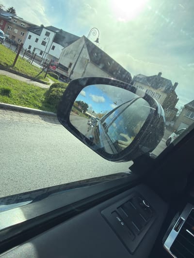The image displays a daytime view from inside a vehicle on a residential street in Reckange-sur-Mess, Luxembourg, under bright, sunny conditions.
The foreground is dominated by the interior of a dark-colored vehicle, showcasing a dusty passenger-side exterior mirror, a section of the dark door panel, and a portion of the dashboard with chrome-accented air vents and various control buttons. The mirror's reflection shows a clear blue sky with white clouds, and the left hand of an individual, likely a passenger or driver, holding a black smartphone. Behind the reflected hand, a road recedes, with a white van and other vehicles visible.
To the left and forward of the vehicle, an asphalt road runs parallel to a paved sidewalk and a grassy area enclosed by a fence constructed from wooden posts and wire. Beyond this, a red pickup truck, partially visible and bearing "COZZARELLI" branding, is situated on the road. A blue rectangular road sign, depicting a left-pointing arrow, is also present. The background features a cluster of diverse residential buildings, some appearing older with distinct architectural styles, while others are more modern, including one with rooftop solar panels. Several black, ornate streetlights with spiral finials are positioned along the street.