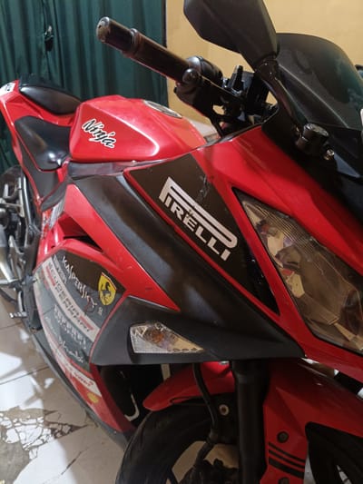 A red and black Kawasaki Ninja sport motorcycle is depicted indoors in the city of Cileungsi, Indonesia. The motorcycle is positioned facing forward and slightly to the left, occupying the majority of the frame. Its bodywork consists of prominent red fairings contrasting with black sections.
Visible branding and decals include a white "Ninja" logo on the fuel tank, a large white "PIRELLI" logo on the upper-right fairing, and a series of stickers on the lower-left fairing: "KASPERSKY," a yellow Ferrari "Prancing Horse" emblem, "SCUDERIA FERRARI," "FERRARI WORLD Abu Dhabi," and a "Shell V-Power" logo.
Key components visible on the motorcycle include the right handlebar with a black grip and a silver screw on the bar end, a dark, dusty windshield, a left rearview mirror, a clear headlight assembly, and a front amber turn signal on the right side. The front wheel and suspension fork are partially visible. The black seat, black frame elements, and a silver footpeg or gear shifter are also discernible on the left side.
The motorcycle exhibits signs of use, including minor scuffs and scratches on its fairings and a dusty windshield. It rests on a light-colored tiled floor, some sections of which show cracks and discoloration. The background features dark green curtains on the left and a plain, light-colored wall further back, indicating an indoor setting, likely a garage or residential space. No individuals are present in the image.