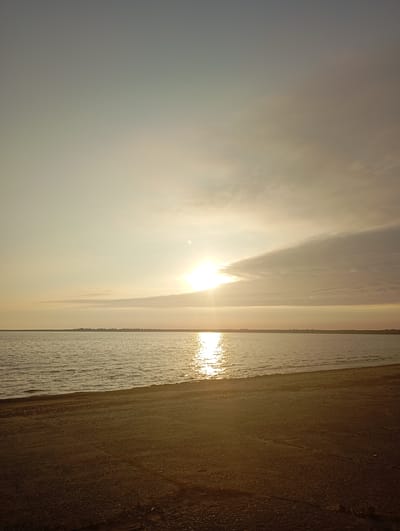 An outdoor, vertical, wide shot shows a beach and a body of water at sunset. The sky is a pale yellow and orange at the horizon, transitioning to a hazy gray at the top of the frame. The sun is a bright yellow orb, partially obscured by a thin, wispy cloud formation to its right. The sun’s reflection creates a shimmering golden path across the water’s surface. The water is calm with gentle ripples. A dark, sandy beach occupies the foreground, with some small pebbles and debris visible. In the distance, along the horizon line, is a faint, silhouetted landmass dotted with what appears to be trees or buildings. This scene is consistent with the coastal landscape of Liepaja, Latvia, specifically a beach area such as Bernati Beach, known for its proximity to the Baltic Sea and its characteristic sandy shores and pine forests in the distance. There are no people or animals visible in the image.