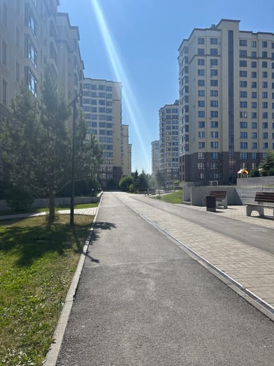 A paved walkway lined with grass and trees runs alongside a series of modern apartment buildings under a clear blue sky. The sun casts long shadows, suggesting it is late afternoon or early morning. The buildings are tall and have many windows, with some balconies visible. There are benches along the walkway, and a small playground structure is visible in the distance. The scene appears to be a residential area, likely a park or courtyard within a housing complex. No people are visible in the image. The weather appears to be sunny and pleasant. The image is taken from a slightly elevated perspective, looking down the walkway.