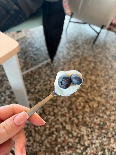 The image displays a hand holding a stainless steel spoon, oriented diagonally from the lower left towards the upper right. The spoon's bowl contains a white, creamy substance topped with two dark blue blueberries. The hand exhibits manicured fingernails with a light pink base and white tips. The background is out of focus, indicating an indoor setting. To the upper left, a light-colored table edge is partially visible. Centered in the mid-ground, a person's lower torso and legs are obscured but visible, dressed in blue denim jeans and dark fabric, positioned near the dark metal frame of a chair. The floor is a speckled, multi-tonal surface, likely terrazzo. This scene takes place indoors in La Clusaz, France.