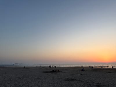 This image captures a serene sunset scene at a beach in Hook of Holland, Netherlands. The sky transitions from a soft blue at the top to warm hues of orange and pink near the horizon, indicating it's either sunrise or sunset. The sun is a vibrant orange orb, partially submerged in the sea.

The foreground is a sandy beach, textured with footprints and scattered debris. Several people are scattered across the beach. Some are walking, while others are gathered in small groups, perhaps socializing or watching the sunset. In the middle ground, a few individuals are walking along the waterline.

In the distance, the sea stretches to meet the horizon, with gentle waves lapping at the shore. A large ferry, displaying the text "Stena Line," is visible on the water, suggesting maritime activity. Several wind turbines are also present in the background, a common sight in the Dutch landscape, particularly near coastal areas and ports.

The overall mood is peaceful and relaxed, typical of an evening at the beach. The soft lighting and the vast expanse of the sky and sea contribute to a sense of tranquility. The presence of people enjoying the natural beauty suggests a pleasant end to the day.