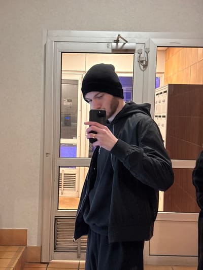 A person in a black beanie and jacket takes a mirror selfie. They are holding a phone with their right hand, the screen facing them. The mirror reflects a room with a window, showing a building with repeating rectangular patterns. The surrounding wall is plain and a light beige color. Tiled flooring can be seen in the bottom-right corner. The window has security features on the sides and top.