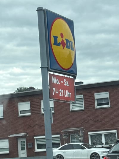 A wide-angle, eye-level, outdoor shot captures a cloudy day in Gronau, Germany, with a focus on a Lidl store's signage and building. The Lidl logo, a circular yellow sign with blue lettering and a red diamond shape, is prominently displayed on a grey pole on the left side of the image. Below the logo, a red rectangular sign reads "Mo.-Sa." and "7-21 Uhr" in white text, indicating the store's operating hours from Monday to Saturday, 7 AM to 9 PM.
The background features a red brick building, presumably the Lidl store, with several windows. Some windows have white rolling shutters pulled down, while others are partially open, revealing patterned blinds inside. A white door with glass panes is visible on the left side of the building. In front of the building, two cars are parked. The car in the foreground is a white sedan, and a portion of another dark-colored vehicle is visible on the right edge of the frame. The sky is overcast with scattered clouds. The street in front of the store is not clearly visible, suggesting the photo may have been taken from a vehicle.