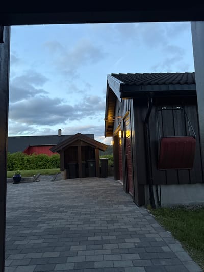 The image depicts a twilight scene in Åmot, Norway, featuring a paved driveway leading to a dark wooden building with red doors and exterior lighting. A red snow shovel is mounted on the wall of the building. To the left, a smaller wooden structure houses trash bins, and further in the background, a red house with a dark roof is visible amidst green foliage. The sky is cloudy and a light blue, indicating the evening. The scene is quiet and serene, with no people present.
