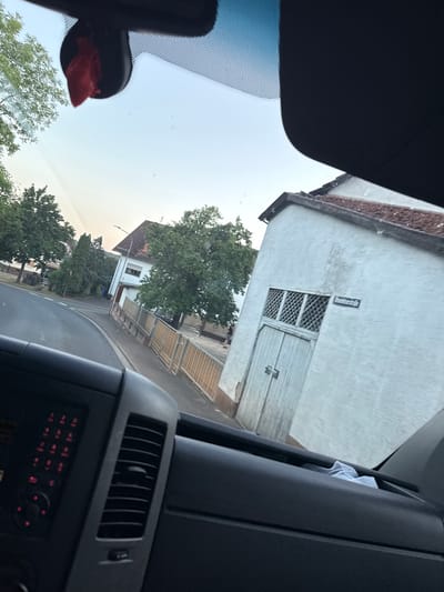 This media file, likely a photograph, depicts a scene viewed from inside a vehicle, looking outwards through the windshield and side window. The perspective suggests the vehicle is parked or moving slowly on a street in Oberwetz, Germany.
The main subjects are the exterior environment and elements of the vehicle's interior. Outside, a weathered, light-colored building with a tiled roof is prominent. It features a large, light-colored wooden door and a smaller, latticed window above it. A rectangular metal sign is affixed to the building's facade. Trees with dense green foliage are visible around the building, suggesting a temperate climate and a season with lush vegetation, likely summer or late spring. In the background, residential buildings and a street can be discerned, along with some fencing.
Interior elements include a dark-colored dashboard with visible air vents and what appear to be control buttons with red indicator lights. A red object, possibly a small accessory or decoration, is attached to a mount on the windshield, near the rearview mirror. A piece of white fabric is tucked into a crevice of the interior paneling.
The time of day appears to be late afternoon or early evening, given the soft, diffused light. The sky is overcast or hazy, indicating fair weather. There are no people visible in the frame. The overall mood is mundane and observational, capturing a slice of everyday life in a residential area. No legible text is discernible, other than the small sign on the building which is too indistinct to read.