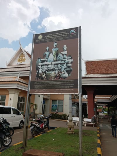 The image shows a large informational billboard in front of a building with a traditional tiled roof and ornate detailing. The billboard displays an image of ancient artifacts, including statues and pottery, and text in Khmer and English. The English text states: "Artifacts are the Precious Heritage of the Nation. We have to Prevent Illegal Trade, Trafficking, Destruction and Loouting of Culturally Important Artifacts." Below this, it details penalties for such offenses and states the information is "Prepared by: Ministry of Culture and Fine Arts" and "Supported by: Mekong-Lancang Cooperation Special Fund".
In the foreground, there are several parked motorcycles and a white van. The environment appears to be an outdoor public area, possibly a government building or cultural institution. The sky is cloudy. People are visible in the background, including someone sitting on a bench and another person walking away from the camera. The overall scene suggests a location in Bavet Quarter, Cambodia, where efforts are being made to protect cultural heritage.