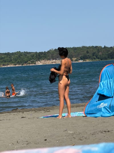 A woman in a black bikini stands on a sandy beach in Atiya, Bulgaria, holding a black garment. She is facing away from the camera, towards the water, where two children are playing. The woman has dark hair pulled up into a bun. In the background, there is a blue beach tent with an orange trim, and a tree-covered hillside rises behind the water. The sky is clear and blue.