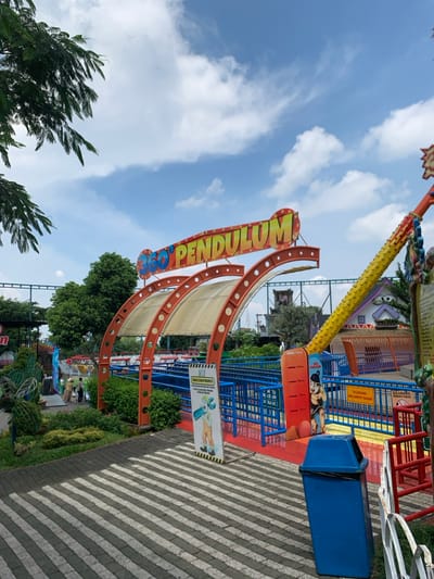 The image shows the entrance to the "360° Pendulum" ride at a theme park in Batu, Indonesia (latitude: -7.885, longitude: 112.525). The entrance is marked with a large orange and yellow sign that reads "360° Pendulum" in bold yellow letters. The entrance is framed by two orange and red arches, each with a white canvas awning. The arches are connected by a metal frame.  There are blue metal railings on either side of the walkway leading to the ride.  A blue trashcan sits in the center of the walkway, directly in front of the ride. There is a poster on the right side of the walkway, warning riders to fasten their seatbelts, and a poster on the left side with instructions for riders to check their height against the height measurement. The surrounding area includes lush green foliage and trees. The sky is a bright blue with fluffy white clouds. A few people can be seen in the background, but none are visible in the foreground.  A colorful amusement ride is visible to the right of the sign, extending away from the camera towards the back of the image.  A small, brightly colored building with a curved roof can also be seen to the right of the sign. The scene is bright and cheerful, suggesting a fun and exciting environment.