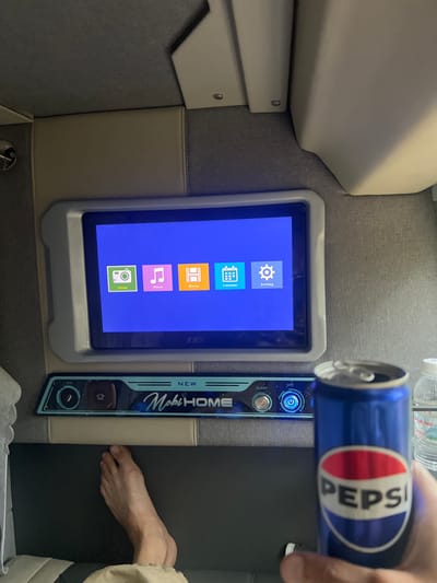 The scene depicts the interior of a private sleeper bus compartment in the city of Aê Thi, Vietnam. A single individual is present, with their right hand holding an open blue Pepsi can in the foreground. Their bare left foot rests on a dark surface in the lower-left, partially covered by light-colored fabric.
Centrally positioned on the wall is a rectangular digital display screen with a gray bezel, currently displaying a blue background with five square application icons: a green camera for "Photo", a pink music note for "Music", an orange film strip for "Movie", a light blue calendar for "Calendar", and a white gear for "Setting." Below the screen, a horizontal control panel features the text "NEW MobiHOME" and "SLEEP," accompanied by illuminated buttons and dials set against a dark, possibly space-themed graphic. To the right of the Pepsi can, a clear plastic bottle of "La Vie" brand water is partially visible. The compartment walls are upholstered in gray fabric and light beige leatherette panels, with similar light beige material covering overhead structural elements.