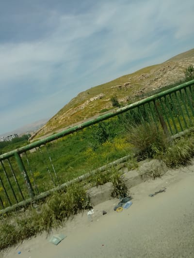 The image shows a hillside landscape taken from what appears to be a roadside or bridge. A green metal railing runs along the lower left portion of the frame, suggesting a barrier. The hillside is covered in vegetation, predominantly grass, with some rocky outcroppings visible. The sky is partially cloudy. There's evidence of litter or debris along the roadside in the foreground. In the distance, some buildings are faintly visible. While it is difficult to verify with certainty, the general features in the image align with areas around Dummar Municipality, Syria, particularly the presence of hillside terrain and sparse vegetation.