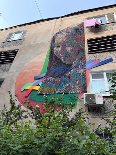 In an urban environment, the side of a multi-story building is adorned with a large mural. The mural features a mosaic portrait of a person's face, composed of numerous small, colorful bottle caps. Abstract shapes and lines in vibrant hues, including pink, orange, yellow, green, blue, and purple, are painted onto the building's facade, creating a dynamic backdrop for the mosaic. A green hose snakes across the mural and down towards the bottom of the frame.
On the building's wall, several windows are visible. One upper-floor window has laundry, including a pink towel and dark clothing, hanging out to dry. Another window on the same level features horizontal blinds. A window on a lower floor is partially obscured by lush green foliage in the foreground. An air conditioning unit is mounted beneath this lower window, bearing the "CHIGO" logo. The building's exterior is a mix of concrete and stone-like textured surfaces. The sky above is a pale blue, with thin overhead wires crossing the frame. The scene is a blend of everyday urban life and artistic expression.