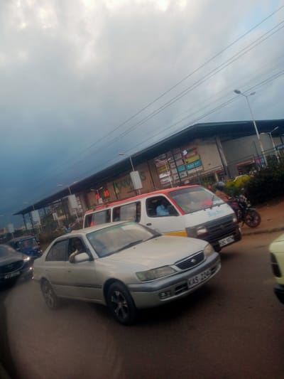 The image captures a street scene in what appears to be Hospital Ward, Kenya, under an overcast sky. A silver sedan is prominent in the foreground, its front end angled towards the viewer. The car displays the license plate "KAS 2204". To its right, a portion of a yellow vehicle is visible. Behind the sedan, a white and red minivan is stopped in traffic, with a driver visible inside. Further back, a blue vehicle, possibly a matatu or tuk-tuk, is also present. A motorcycle is parked near the minivan. Dominating the background is a long building with a dark roof and a facade featuring various signs, including "ANAS" and what appears to be a supermarket or shopping center. The street itself is paved, and the overall impression is of moderate traffic congestion. The lighting suggests it is daytime, but the cloudy conditions contribute to a subdued atmosphere.