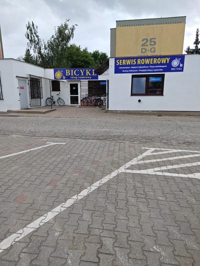 The image shows a bicycle shop and repair service called "BICYL SKLEP ROWEROWY" and "SERWIS ROWEROWY" located at Ulica Jana III 25 D-G in Poznan, Poland. The building has a white facade with a blue sign above the entrance. A green bicycle is parked outside the shop, and several other bicycles are visible inside and in a bike rack. A window on the building displays a "SHIMANO" logo. The shop is situated in an urban environment with a paved parking lot in front, marked with white lines. The sky is overcast.